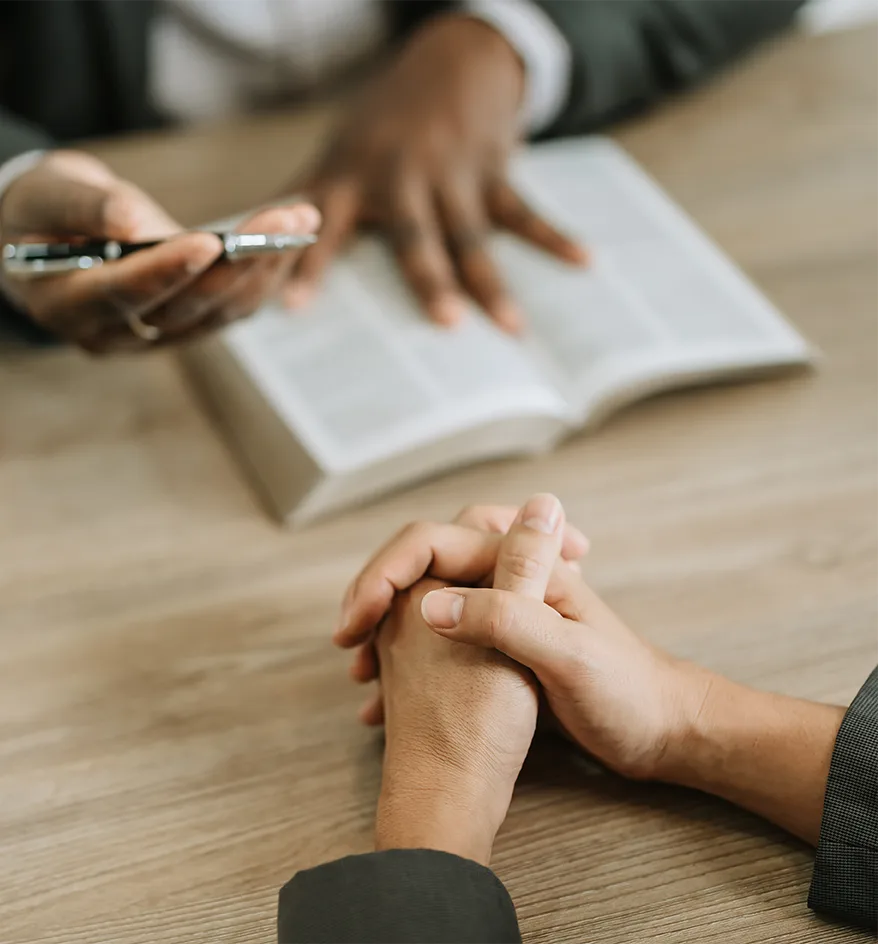 A representative with a pen and an open binder talks across a table with a client whose hands are clasped