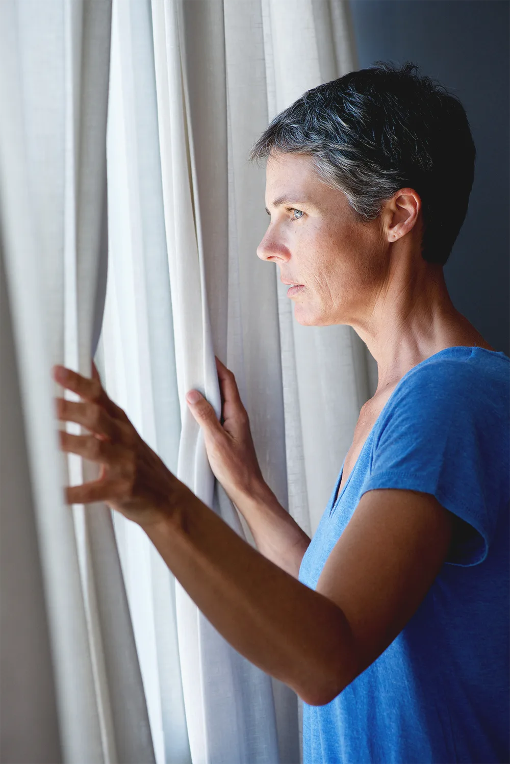 A woman in her 50s with short gray hair looks out a window, holding the curtain