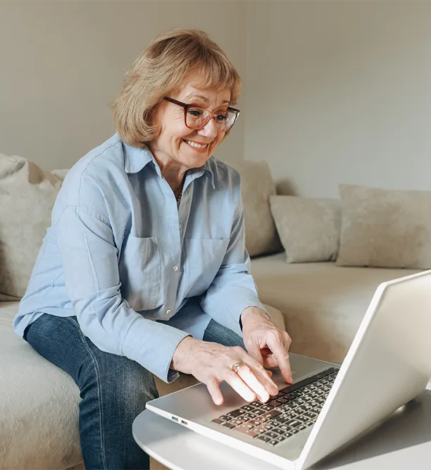 A smiling woman in her early 60s with glasses works on a laptop from her sofa