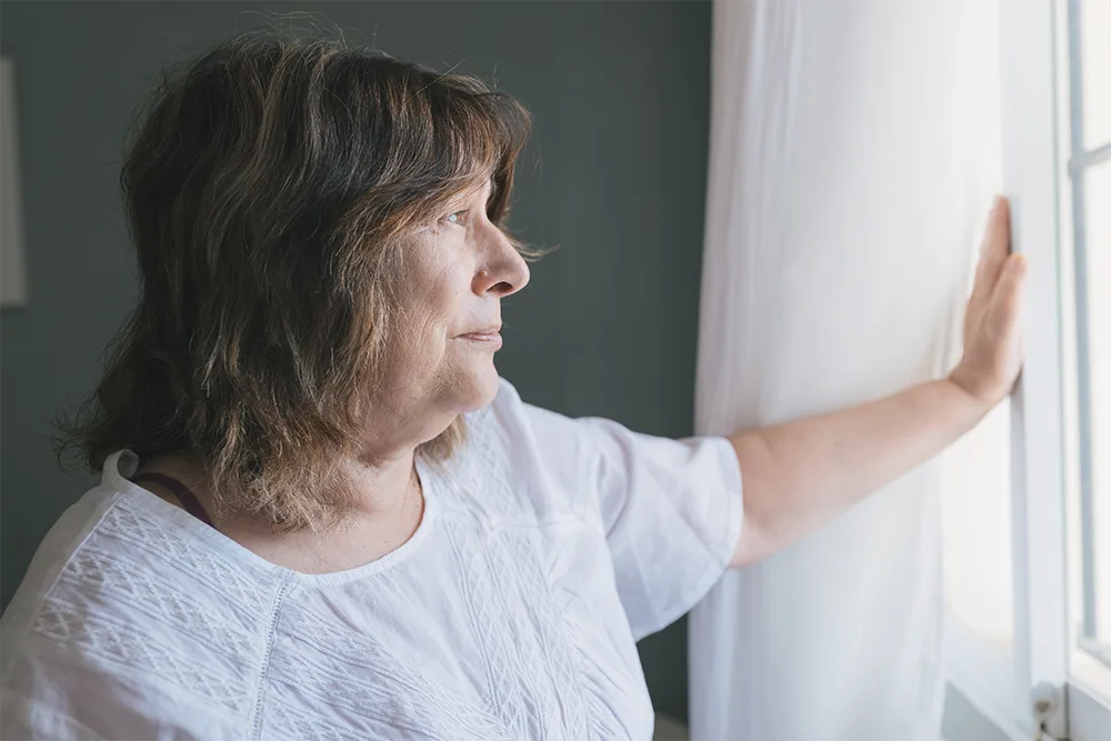 A woman in her late 50s looks out a window, one hand resting on the curtain