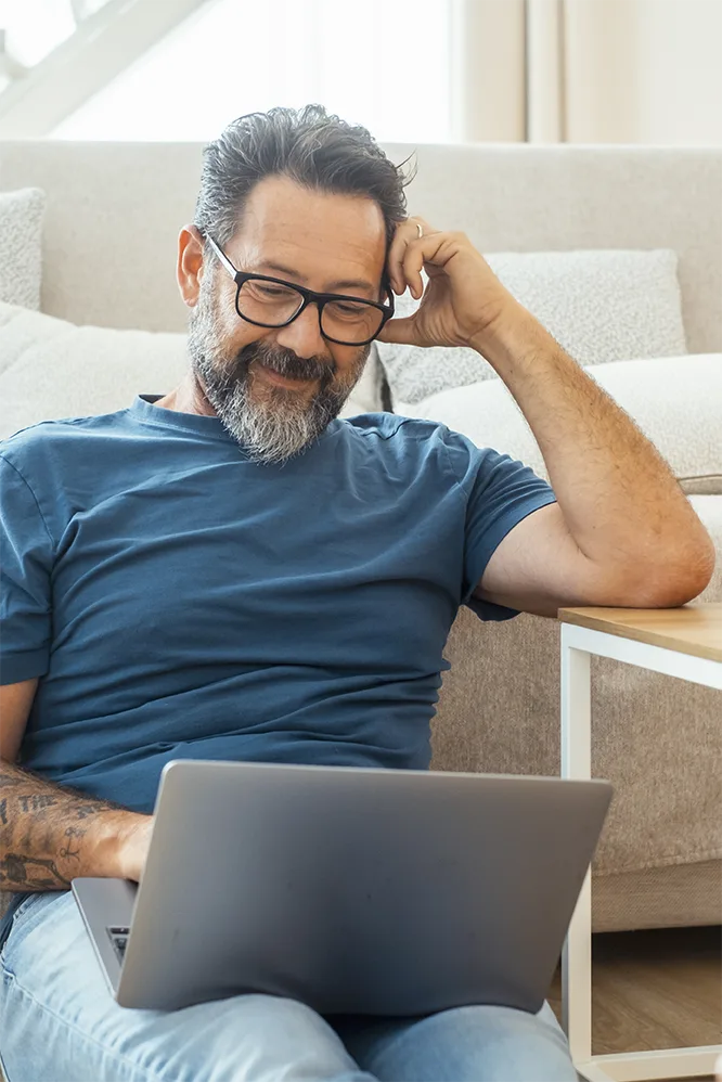 A man in his 50s with glasses and a gray beard smiles at his laptop while sitting by the sofa